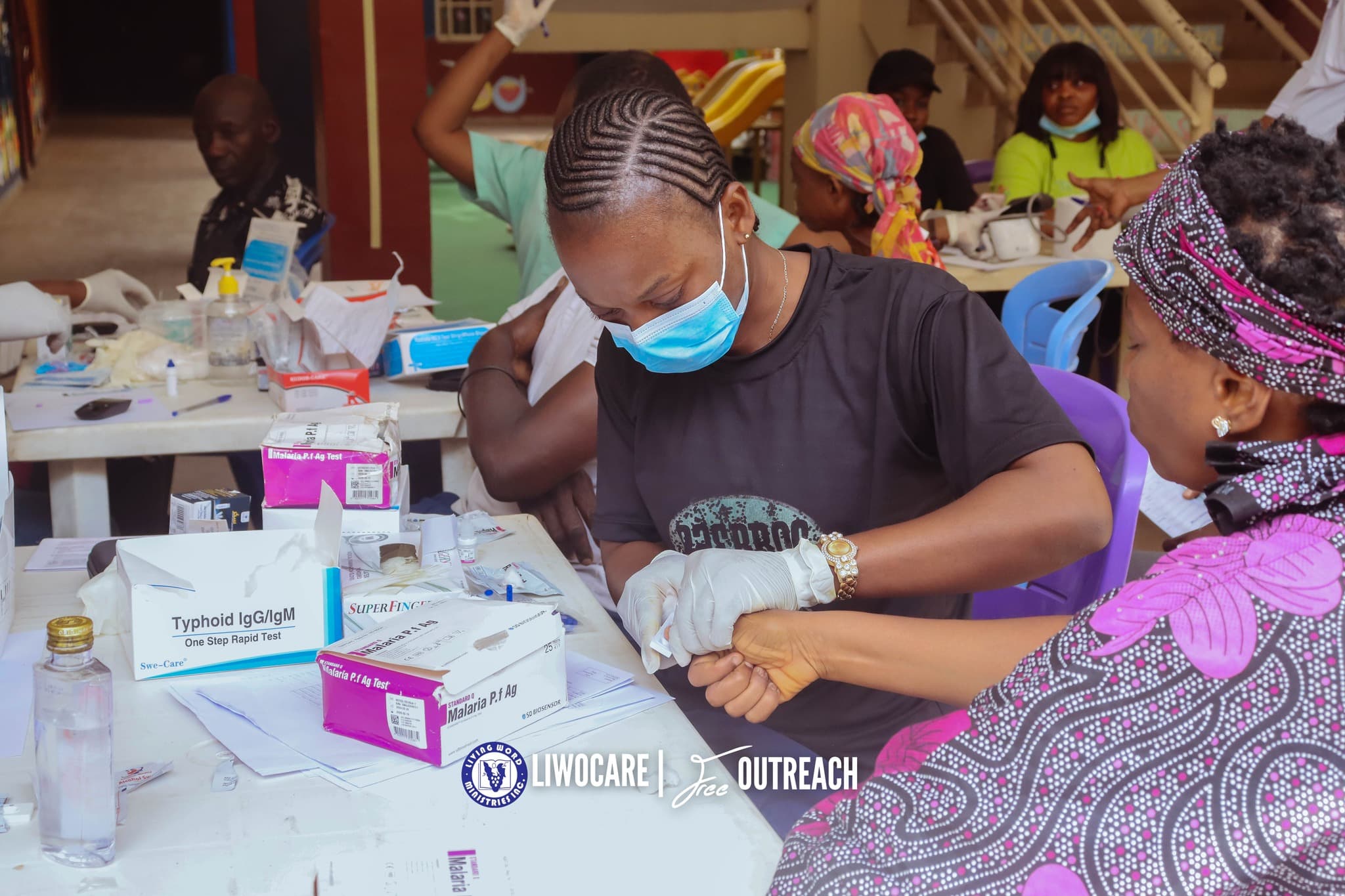 A medical professional wearing a mask and gloves performs a rapid diagnostic blood test for a patient at a table labeled for Typhoid and Malaria testing.