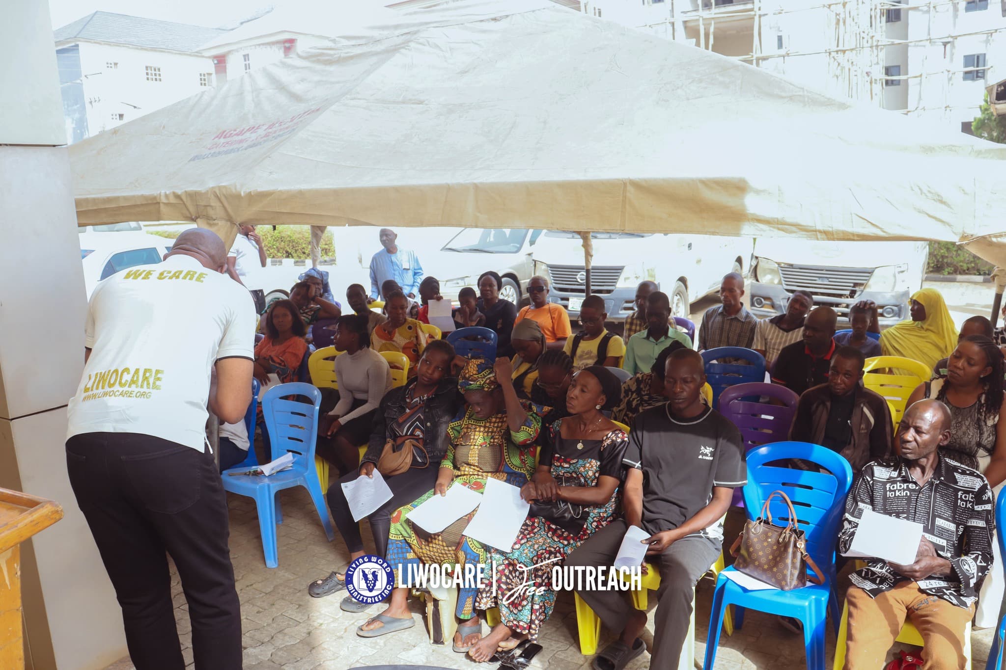 A man in a white "LIWOCARE" shirt speaks to a large group of people seated under a tan canopy outdoors.