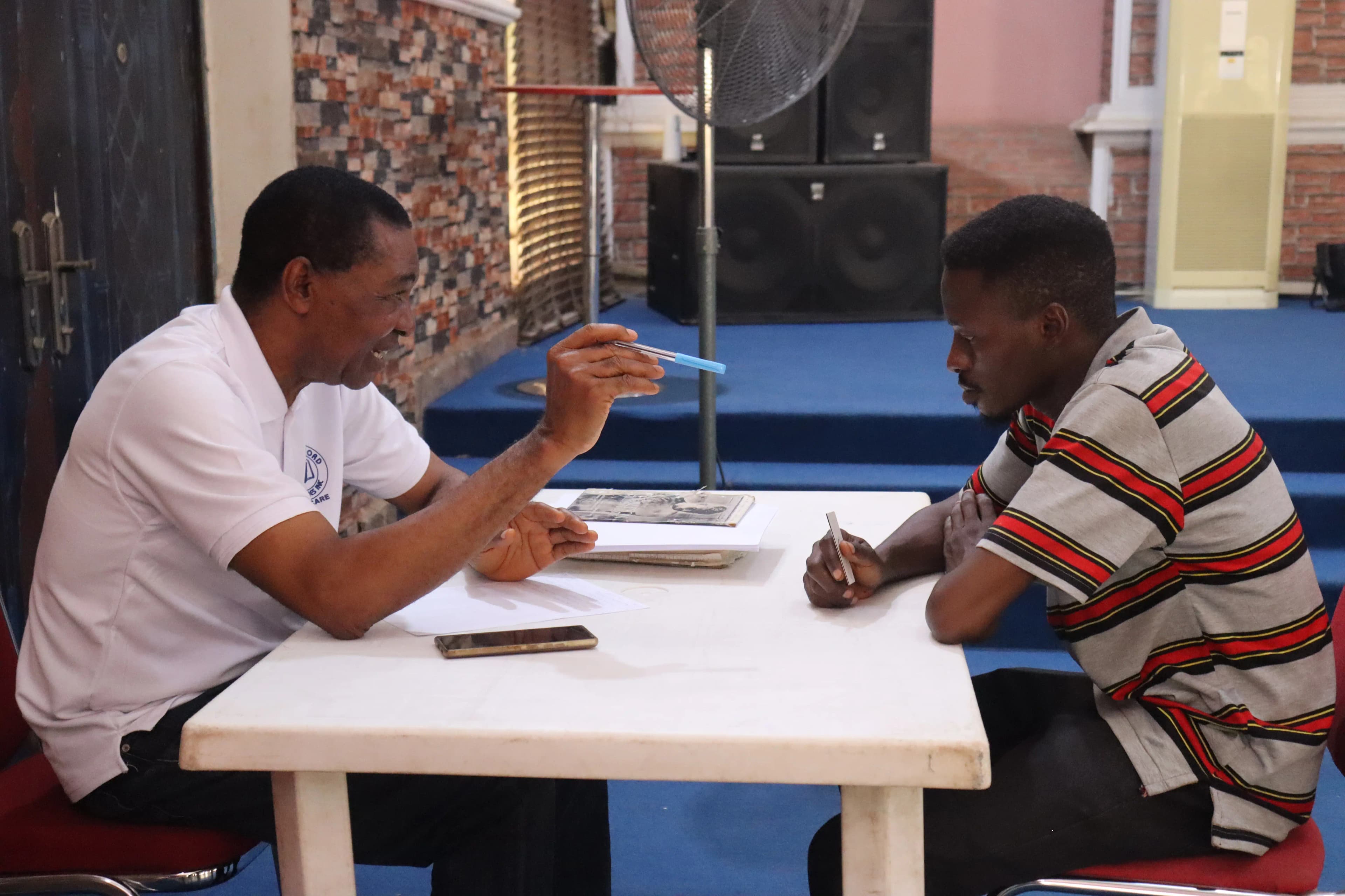 A man in a white polo shirt sits across a table from a young man