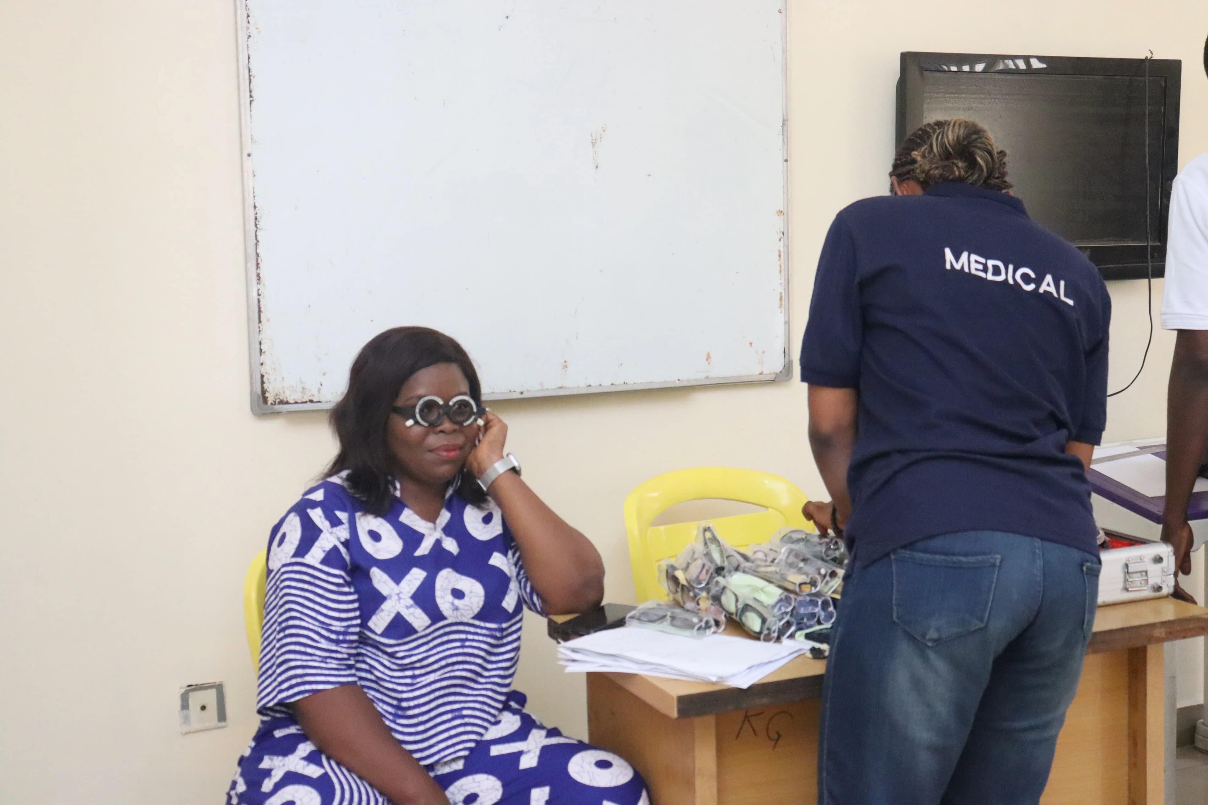 A woman in a blue and white patterned outfit wears trial lens frames during an eye exam, while a medical worker in a dark blue shirt stands nearby.