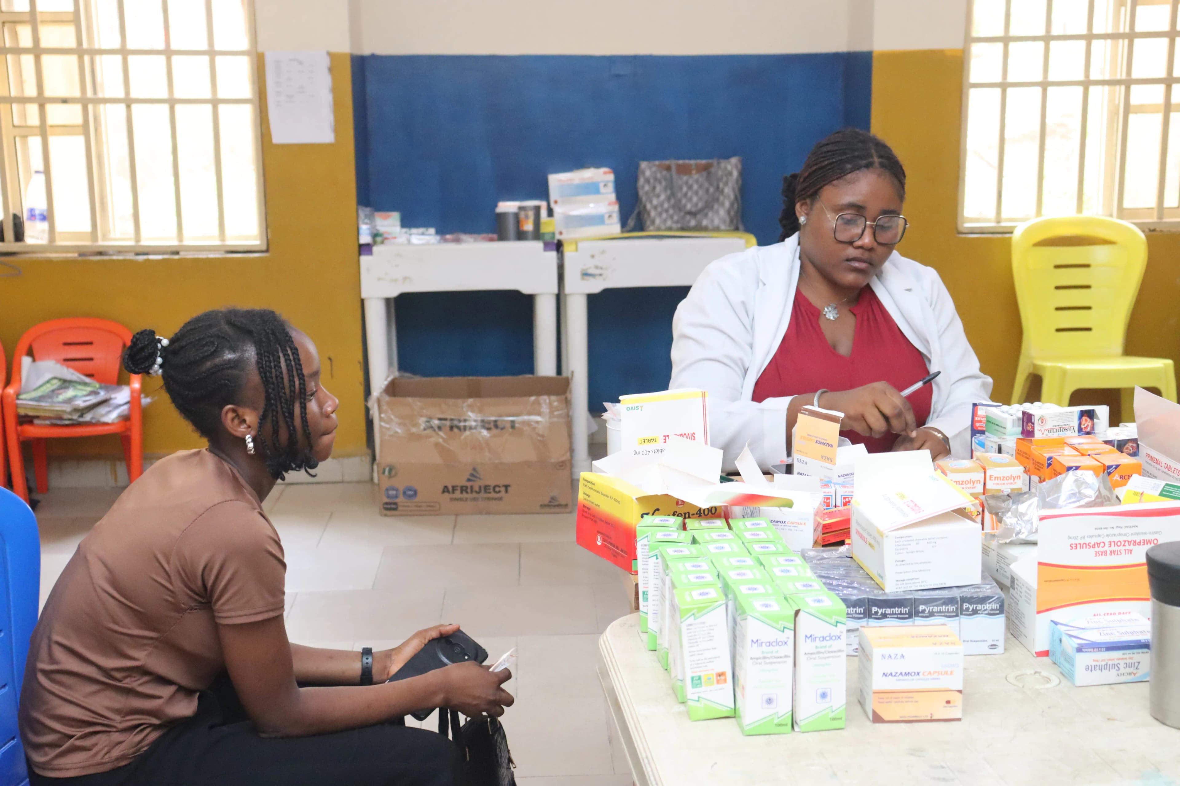 A pharmacist in a white lab coat organizes various boxes of medication on a table while a young woman sits waiting in front of her.