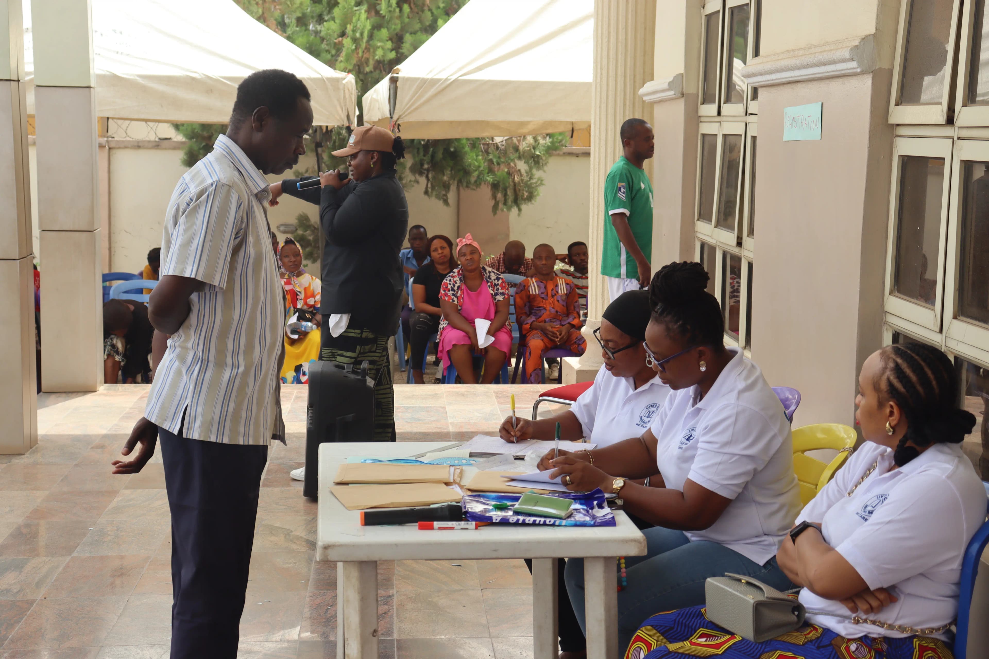 A man standing in front of 3 women seated, while 2 women are writing on paper with people in the background