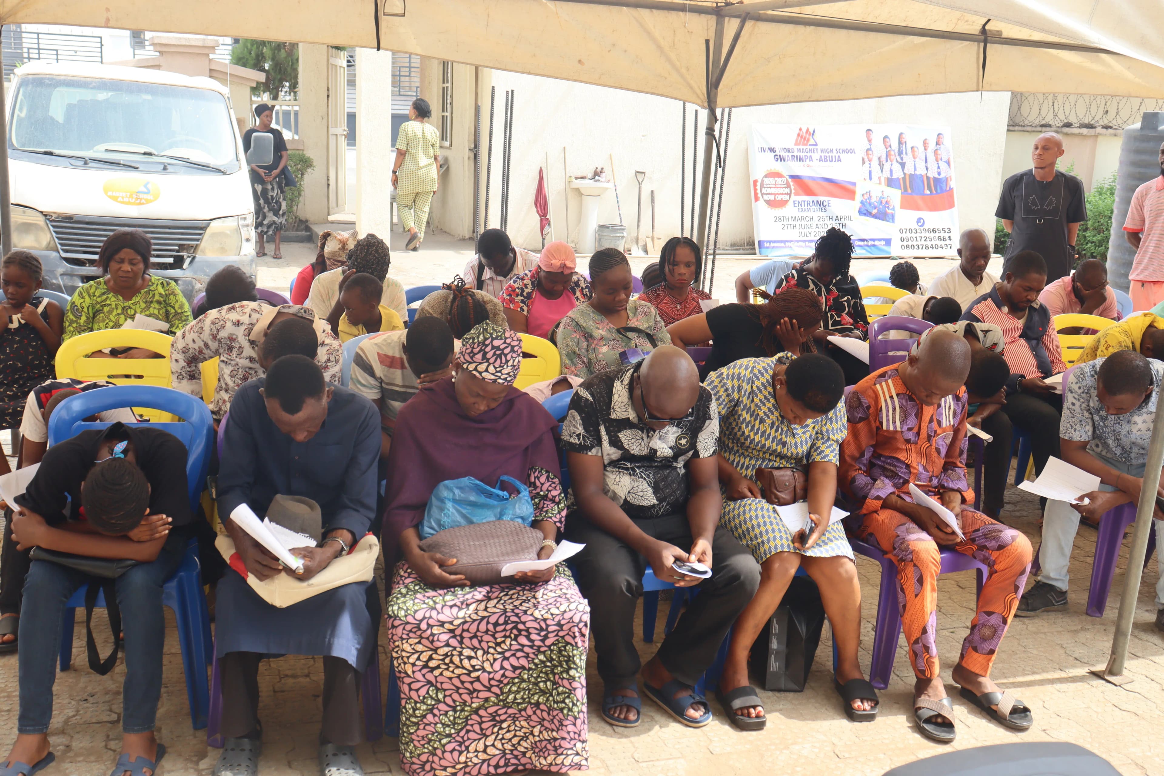 A crowd of people sitting in chairs under a canopy with heads seemingly bowed