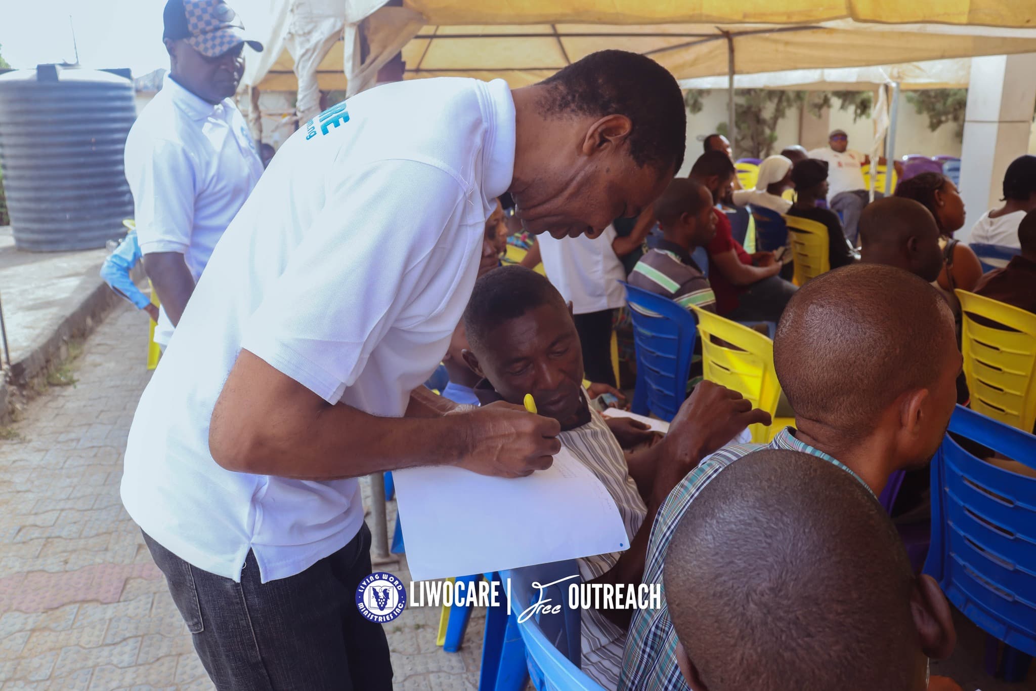 A man bending over while standing and writing on a notepad, seemingly talking to a young man seated amongst people